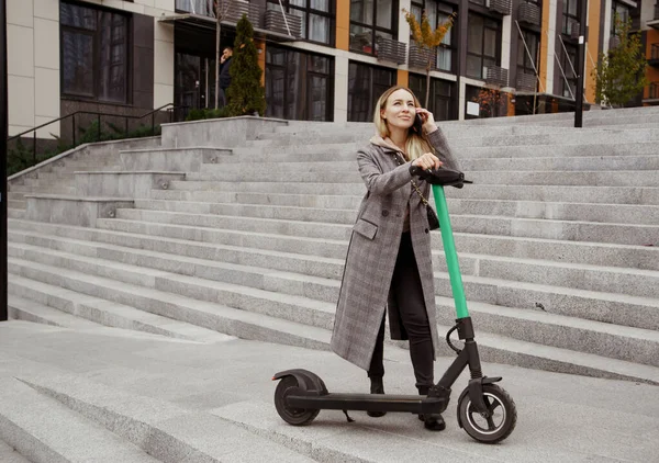 Mujer feliz de pie cerca de scooter eléctrico, mirando al cielo y hablando por teléfono con un amigo. Mujer joven en abrigo acogedor contento de que hay una oportunidad de alquilar un vehículo eléctrico. — Foto de Stock