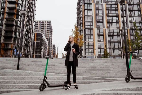 Hij huurde scooter. Herfst in de grote stad. Man in casual kleding bespreekt voorwaarden van het huren van scooter door zijn telefoon. Appartement blokken op de achtergrond. Elektrische scooters voor publiek gebruik buiten. — Stockfoto