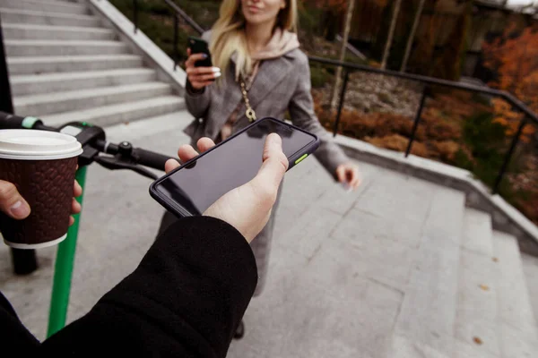 Unusual romantic date on electric scooters. First person view. Man holding smartphone with blank copy space screen, cup of coffee and wheel. Woman standing near with phone in her hand and smiling. — Stock Photo, Image