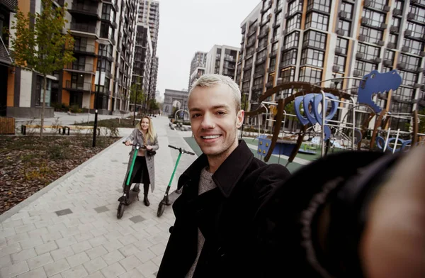 Lykkelige unge par har en god tid ridning e-scootere. Hurtig tur koncept. Romantisk date. Ung blond mand holder kameraet og tager selfie med sin attraktive kæreste. - Stock-foto