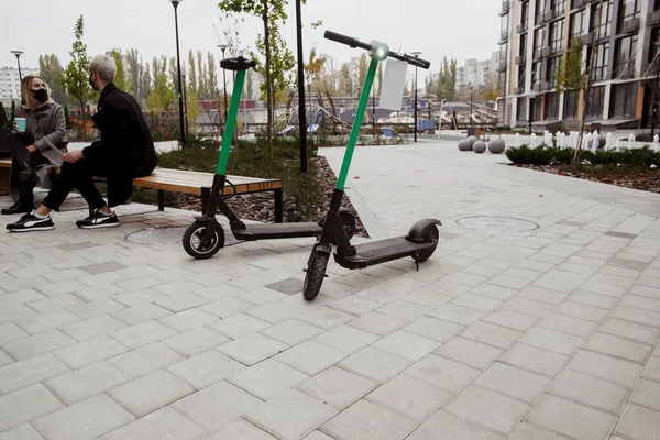 Concéntrate en dos scooters eléctricos. Hombre y mujer en segundo plano sentados en bancos, pasando tiempo juntos y hablando de algo interesante. Manera cómoda de viajar por la ciudad. — Foto de Stock