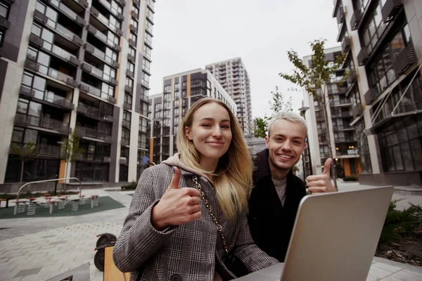 Young man and woman like working outside. Couple sitting on bench and thumbing up. Modern apartment blocks on background. Young attractive woman and handsome man smiling and looking at camera.