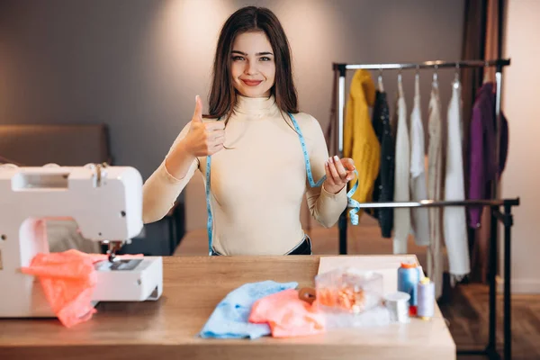 Mujer joven modista con máquina de coser. Costurera está sosteniendo medida y da pulgar hacia arriba en el taller. Creación de cursos de diseño de ropa en línea. — Foto de Stock