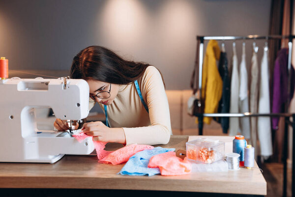 Красивая портниха в очках шьет одежду на швейной машинке. Швея в мастерской. Создание онлайн курсов по дизайну одежды.