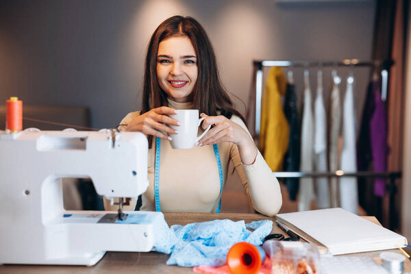 Молодая портниха шьет одежду на швейной машинке. Улыбаясь и пить кофе чай швея в мастерской. Создание онлайн курсов по дизайну одежды.