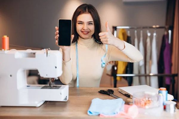 Mujer modista joven cose ropa en la máquina de coser. La costurera muestra felizmente su teléfono en el taller. Creación de cursos de diseño de ropa en línea. — Foto de Stock