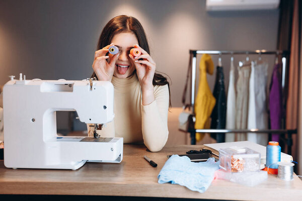 Young dressmaker woman with sewing machine. Seamstress covering eyes with threads in workshop. Creating online clothing design courses.