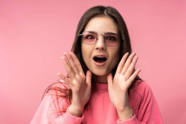 Gözlüklü çekici genç bir kadının fotoğrafı, sevimli ve şok olmuş bir yüz. Sıradan pembe tişört giyer. Beyaz pantolon. İzole edilmiş pembe arka plan.