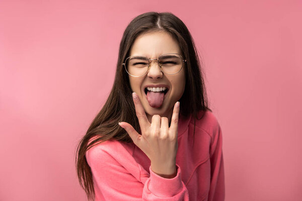 Photo of attractive young woman in glasses gesture rock-and-roll, wears casual pink t-shirt white pants isolated pink color background