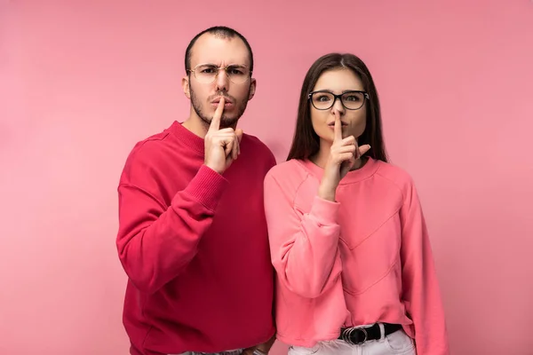 Photo of sweet couple in glasses stands and shows shhh. Male and female look serious and strict, isolated over pink background — Stock Photo, Image