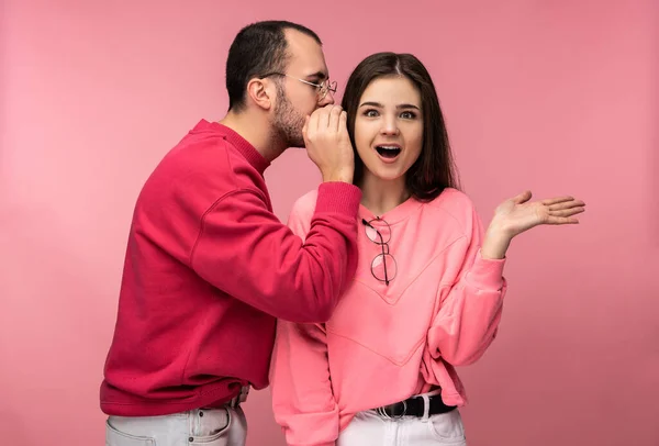 Photo of sweet couple, man whispers some secrets or sales to his girlfriend and she is surprised, isolated over pink background — Stock Photo, Image