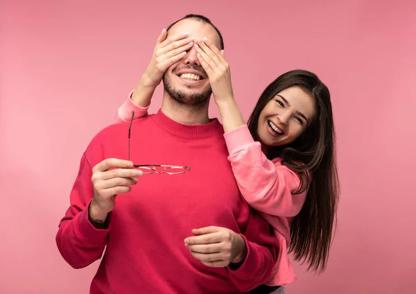 Photo of sweet couple, woman behind her boyfriend back closes his eyes up. Male and female are in love look blessed and smile, isolated over pink background — Stock Photo, Image