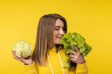 Çekici kadının elinde lahana, salata ve sağlıklı yemek kokusu var. Sıradan sarı kapüşonlu, izole sarı arka plan giyiyor.