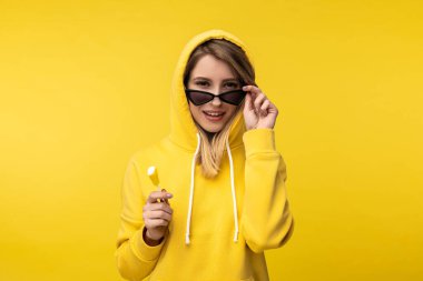 Lolipoplu hoş bir bayanın fotoğrafı gözlüğünü tutuyor ve gülümsüyor. Sıradan sarı süveter giyiyor, izole edilmiş sarı arka plan