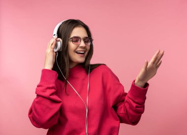 Gözlüklü çekici bir kadının fotoğrafı müzik dinler, dans etmeyi sever. Sıradan pembe süveter giyiyor. Beyaz pantolon, izole pembe arka plan.
