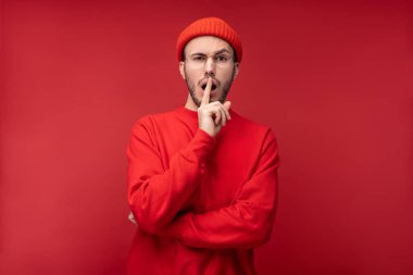 Sakallı, gözlüklü ve kırmızı elbiseli çekici bir adamın fotoğrafı. Erkek şokta ve meraklı, kırmızı arka planda izole edilmiş.