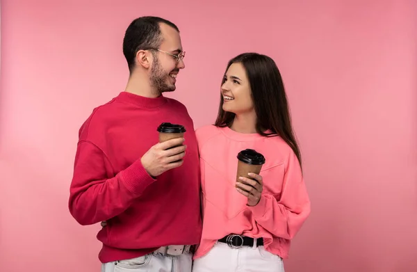 Photo of attractive man wih beard in red clothing and woman in pink holds coffee and gawp on each other. Couple looks happy, isolated over pink background — Stock Photo, Image