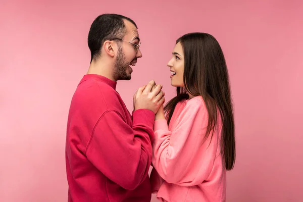 Фото привлекательного мужчины с бородой в красной одежде и женщины в розовых объятиях за руки друг друга с открытым ртом. Пара выглядит счастливой, изолированной на розовом фоне — стоковое фото