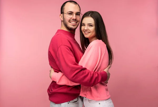 Foto de homem atraente wih barba em roupas vermelhas e mulher em rosa abraçar uns aos outros e sorrir. Casal parece feliz, isolado sobre fundo rosa — Fotografia de Stock