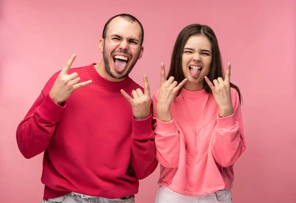 Photo of attractive man wih beard in red clothing and woman in pink show gesture rock-and-roll with tongue sticking out, isolated over pink background — Stock Photo, Image