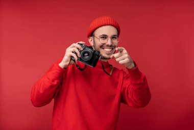 Sakallı, gözlüklü ve kırmızı elbiseli çekici bir adamın fotoğrafı. Mutlu adam kamerayı tutuyor ve güzel görünüyor, kırmızı arka planda izole edilmiş.