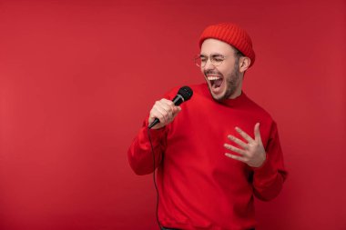 Sakallı, gözlüklü ve kırmızı elbiseli çekici bir adamın fotoğrafı. Erkek mikrofon karaokesinde şarkı söylüyor, kırmızı arka planda izole edilmiş.