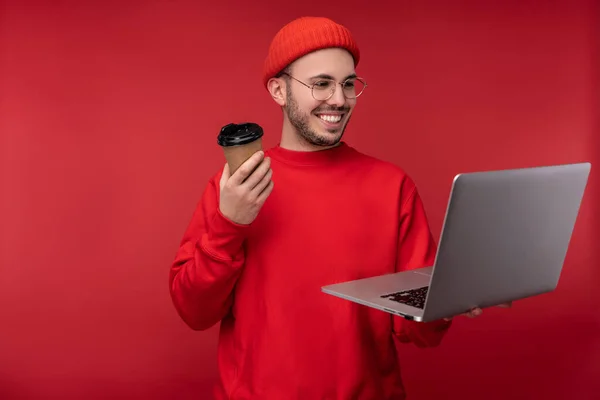 Foto van een aantrekkelijke man met baard in bril en rode kleding. Leuke man houdt laptop en koffie, geïsoleerd over rode achtergrond — Stockfoto