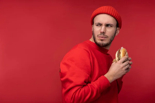 Foto van een aantrekkelijke man met baard in bril en rode kleding. Man houdt hamburger en probeert te verbergen, geïsoleerd over rode achtergrond — Stockfoto