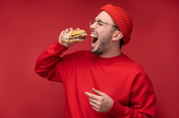 Sakallı, gözlüklü ve kırmızı elbiseli çekici bir adamın fotoğrafı. Erkek hamburgeri yer ve yer, kırmızı arka planda izole olur. — Stok fotoğraf