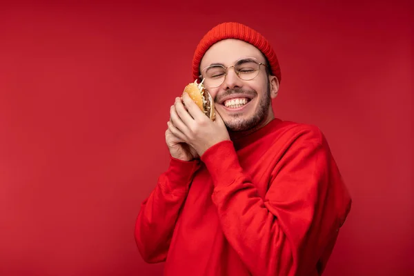 Foto van een aantrekkelijke man met baard in bril en rode kleding. Mannelijke schattige knuffels hamburger en glimlacht, geïsoleerd over rode achtergrond — Stockfoto