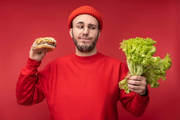 Foto van een aantrekkelijke man met baard in bril en rode kleding. Man houdt salade en hamburger, neemt de beslissing tussen gezond en junk food, geïsoleerd over rode achtergrond — Stockfoto