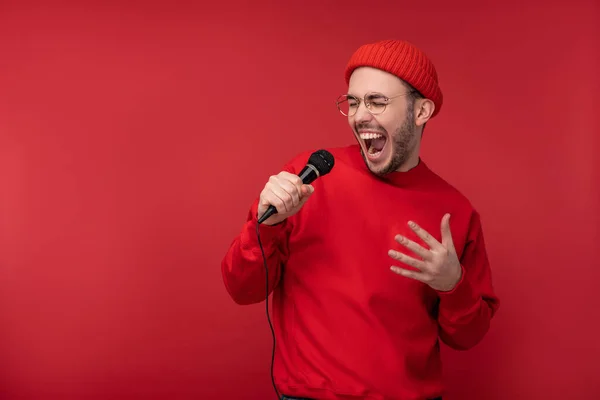 Sakallı, gözlüklü ve kırmızı elbiseli çekici bir adamın fotoğrafı. Erkek mikrofon karaokesinde şarkı söylüyor, kırmızı arka planda izole edilmiş.