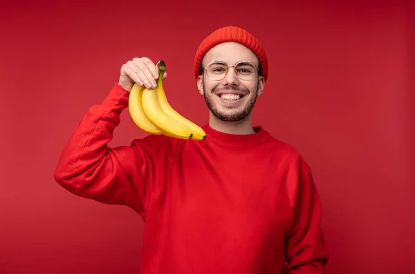 Foto van een vrolijke man met baard in bril en rode kleding. Houdt bananen en glimlachen, gezonde levensstijl, geïsoleerd over rode achtergrond — Stockfoto