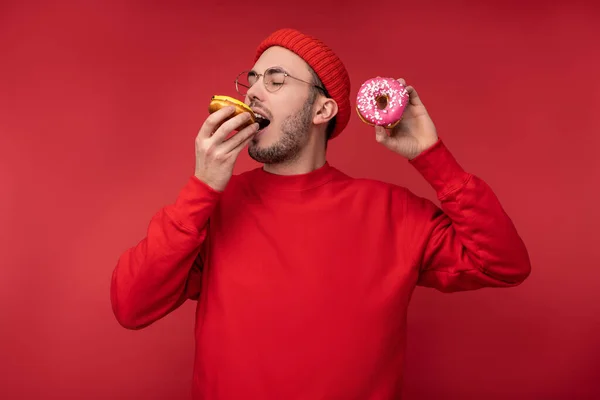 Foto van een vrolijke man met baard in bril en rode kleding. Geniet van het eten van zoetigheden donuts, geïsoleerd over rode achtergrond — Stockfoto