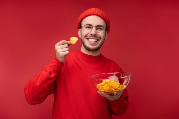 Gözlüklü ve kırmızı elbiseli mutlu adamın fotoğrafı. Kırmızı arka planda izole edilmiş bir tabak cipsi tutar ve yer. — Stok fotoğraf