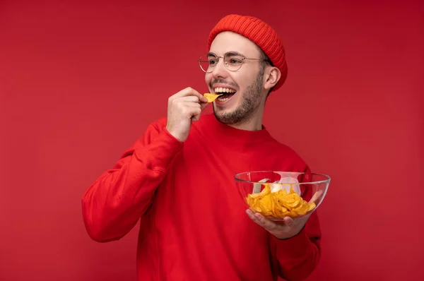 Foto van een vrolijke man met baard in bril en rode kleding. Houdt en eet een plaat chips, geïsoleerd over rode achtergrond — Stockfoto