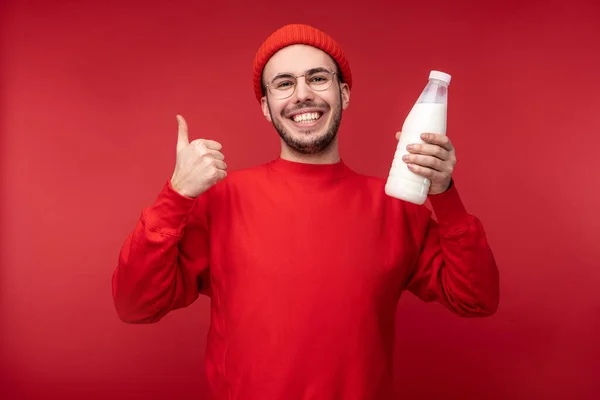 Kırmızı elbiseli mutlu bir adamın fotoğrafı süt ürünleri bulundurur. Kırmızı arka planda izole edilmiş bir şekilde başparmağını kaldırır. — Stok fotoğraf