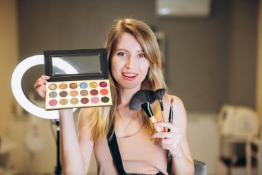 Sarışın makyaj sanatçısı fırça ve göz farı paleti gösteriyor. Güzel bir kadın gülümser ve makyaj ve göz farı için fırça tutar.