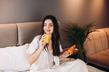 Kadın yatakta portakal suyu içiyor. Beyaz pijamalı bir model oturur ve elinde bir bardak portakal suyu taşır. Genç bir kadın için sabahları yatakta kahvaltı