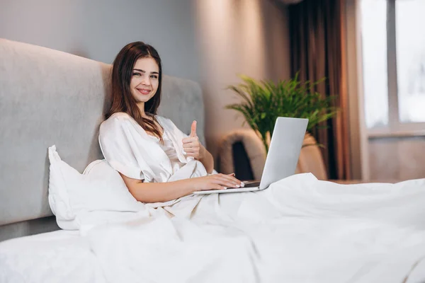 Young beautiful woman with laptop in bed. A brunette with a laptop looks at the camera and shows cool. A woman works at a computer in her bedroom at home