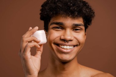 Yakından bakınca piercingli çekici siyahi adamın yüzü için beslenme kremi tutuyormuş. Çıplak gövde, izole edilmiş kahverengi arkaplan