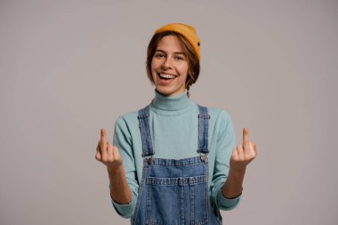 Tatlı bayan hipster 'ın fotoğrafı eğleniyor ve orta parmakları gülümseyerek gösteriyor. Beyaz kadın kot pantolon giyer ve gri renkli şapka giyer.