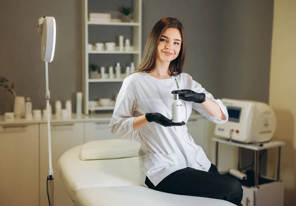 Kozmetoloji kliniğinde danışmanlık. Beyaz önlüklü ve siyah eldivenli kadın güzellik doktoru bir şişe krema gösteriyor. Sağlık hizmetleri kavramı.