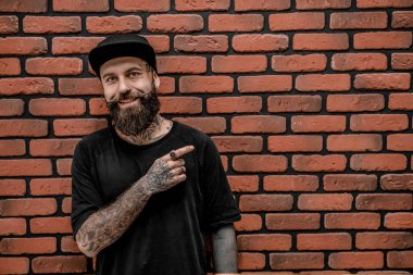 Tişört ve şapka giyen yakışıklı, eski moda hippi, poz ver ve gülümse. Tuğla bir arka planda.