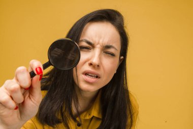 Sevimli bir kadının elinde büyüteçle bir şey aradığı fotoğraf. Sarı gömlek giyiyor, izole edilmiş sarı arkaplan