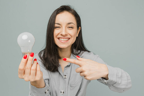 Фото милой улыбчивой женщины держит лампу и указывает на нее. Носит серые платья, изолированный серый цвет фона