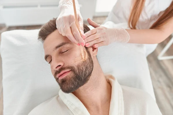 Kozmetik ve yüz bakımı kavramı. Bir kadın güzellik uzmanı, epilasyon yaptıran bir adam için yüz ve sakal modelliği yapıyor. Balmumuyla deplasyon