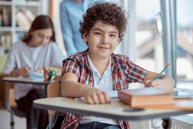 Okulda masa başında oturan kıvırcık çocuk yazı sınavı yapıyor. Masalarda oturan ilkokul öğrencileri.