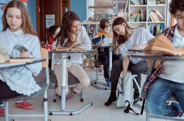 İlkokul çocukları sıralarında oturup sınıfta kitap okuyorlar. Sırt çantasında kitabını arayan genç bir kız.