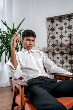 Genç siyahi bir adam, arka planda bir sandalyede poz veriyor. Beyaz gömlekli şık afro erkek manken bir restoranda veya stüdyoda modern bir iç mekanda poz veriyor..